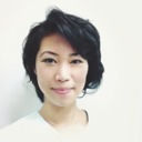 A person with short, dark hair is smiling gently against a plain white background. They are wearing a light top, and the image is softly lit, highlighting their calm expression.