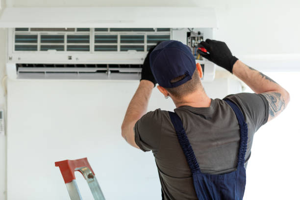 A technician in a cap and gloves stands on a ladder, diligently repairing a wall-mounted air conditioning unit as part of Indianapolis property management. His back faces the camera while he skillfully uses a screwdriver on the unit.