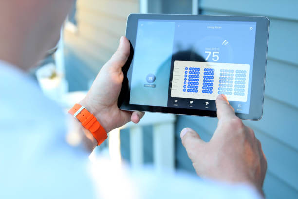A person with an orange wristband is holding a tablet displaying a smart home interface. The screen shows a thermostat set to 75 degrees and a grid of circular controls. The background features the side of a house.