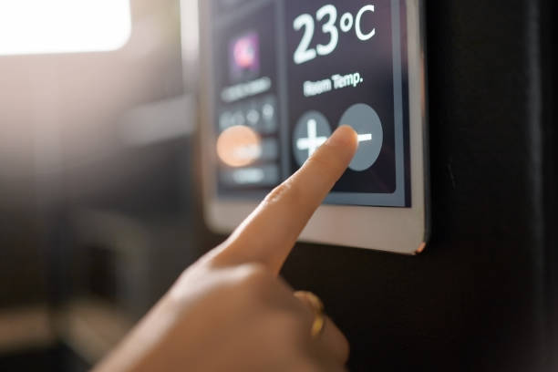 A person adjusting the temperature on a touchscreen thermostat set to 23°C. The screen displays controls labeled with plus and minus signs. The environment appears dimly lit, and the focus is on the finger pressing the plus button.