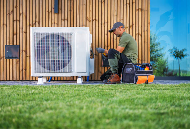 A technician in a cap and work attire kneels on the grass, expertly servicing an outdoor HVAC unit near a wooden building wall. A tool bag is open beside him, while reflections of trees dance across the window, highlighting the harmony of nature and property management.