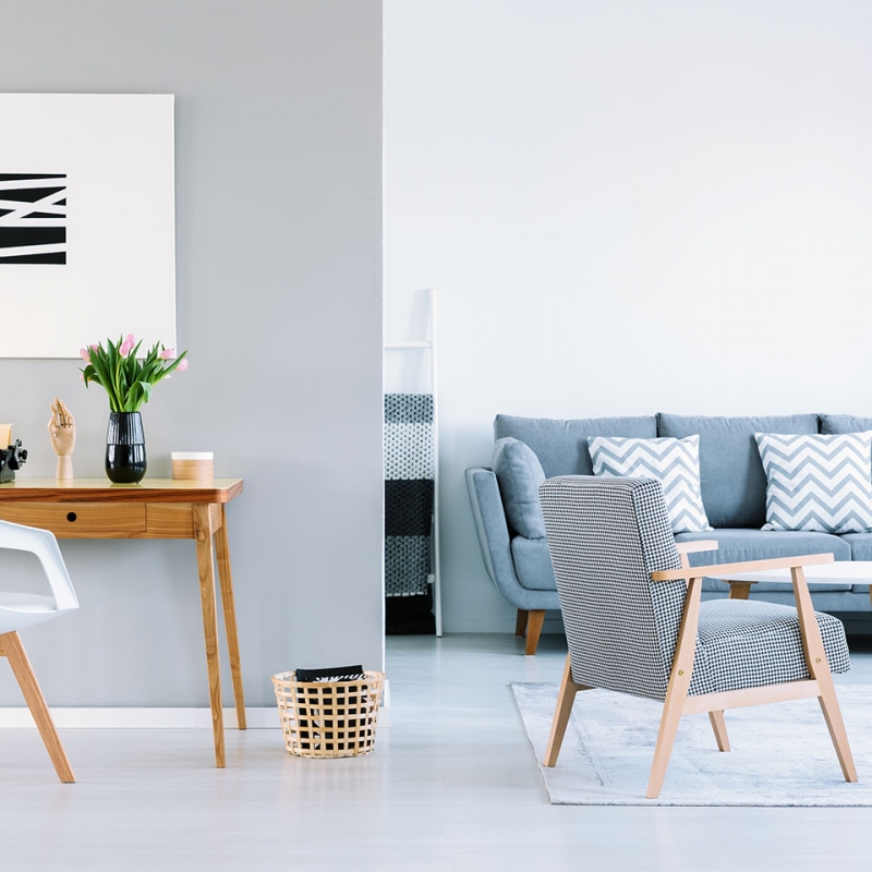 Minimalist living room with a light gray sofa, patterned cushions, a wooden armchair, and a sleek white desk. A black and white abstract painting hangs on the wall.
