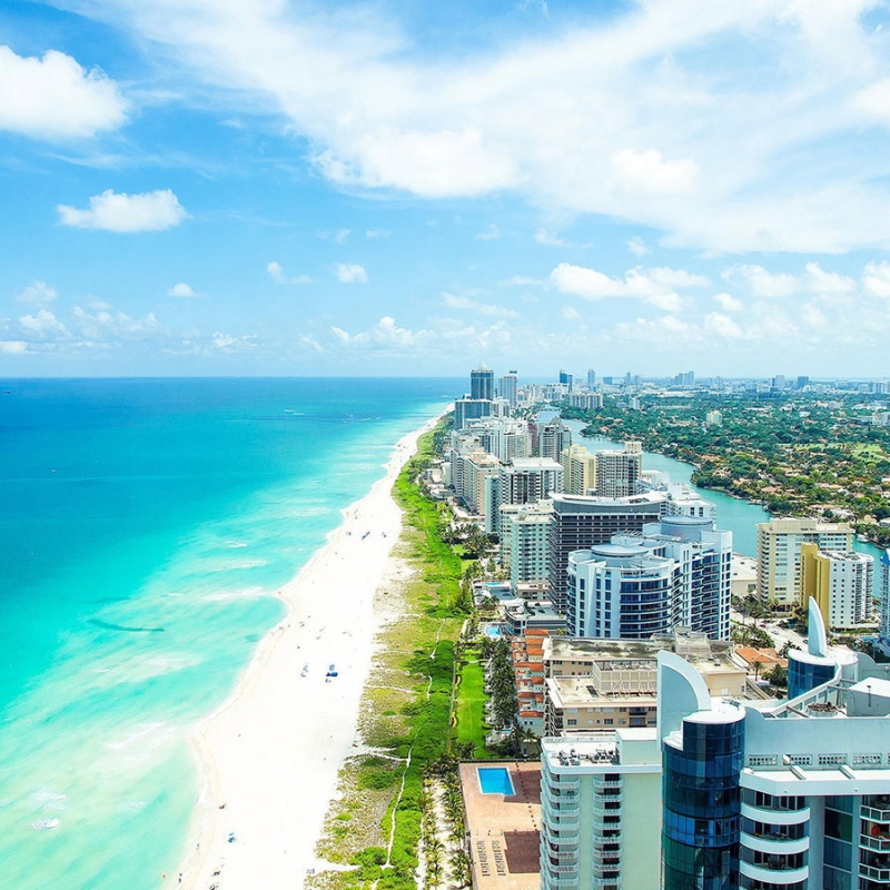 Aerial view of a coastal cityscape with high-rise buildings along a sandy beach. The turquoise ocean stretches to the horizon under a partly cloudy blue sky. Lush greenery peeks through the urban area.